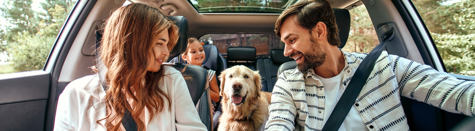 a couple and their young female child smile at a Golden Retriever while sitting inside their SUV 
